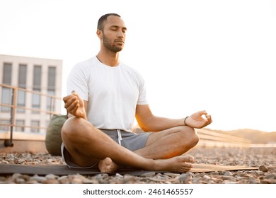 Handsome male meditating in lotus position while sitting on mat on roof of modern house. Young athletic man doing yoga at sunset on urban background. - Powered by Shutterstock