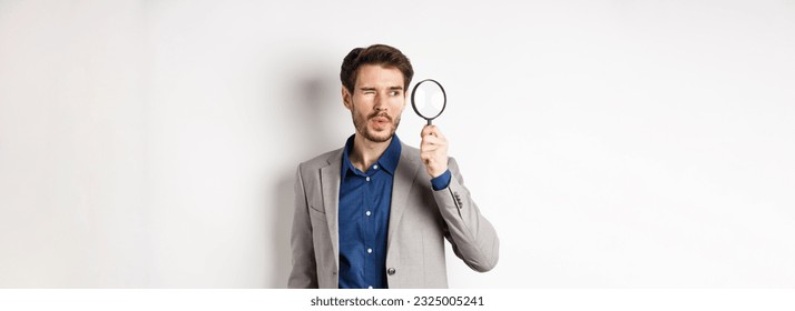Handsome male model in suit looking through magnifying glass with interest, seeing something aside, white background. - Shutterstock ID 2325005241
