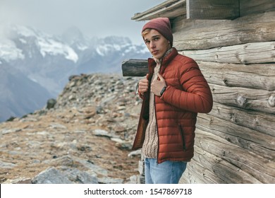 Handsome Male Model Wearing Warm Sweater, Winter Coat And Hat Posing Over Mountains With Snow. Active Fashion Clothes For Cold Weather.