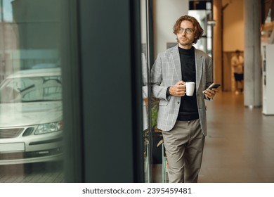 Handsome male office worker use mobile phone and drink coffee while standing near window - Shutterstock ID 2395459481