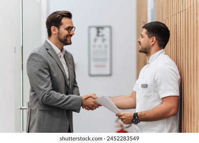 Handsome male patient standing and talking with good looking male doctor in a hall of modern clinic for diagnosis and treatment of eye and sight diseases. Healthcare and medical concept. - Powered by Shutterstock