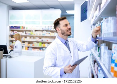 Handsome Male Pharmacist In White Coat Working In Pharmacy Store Or Drugstore. Checking Medicines On His Tablet Computer. Healthcare And Apothecary.