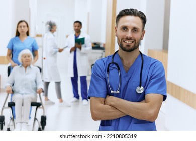 Handsome Male Physician With Stethoscope Standing In Front View Of Hospital Lobby With Doctors In White Coats, Medical Staff And Patients. Working Day In Medical Clinic