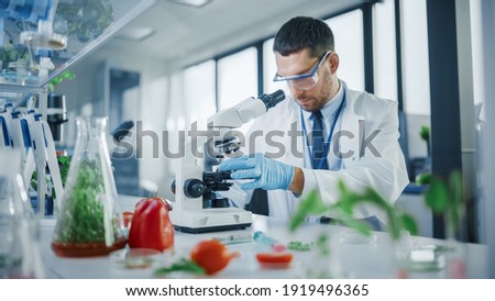 Handsome Male Scientist in Safety Glasses Analyzing a Lab-Grown Food Sample Through an Advanced Microscope. Microbiologist Working on Molecule Samples in Modern Laboratory with Technological Equipment