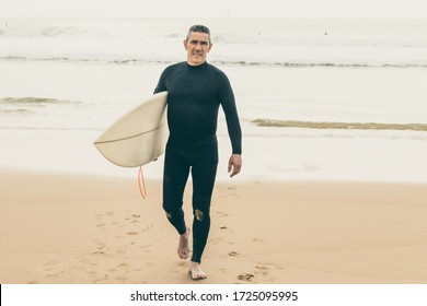 Handsome male surfer looking at camera. Full length view of middle aged man in wetsuit holding surfboard and walking on sandy sea coast. Surfing concept - Powered by Shutterstock