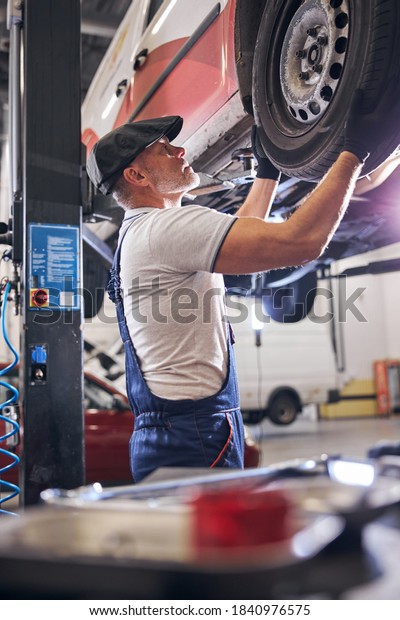 Handsome male technician in cap working with\
lifted car in automobile repair\
garage