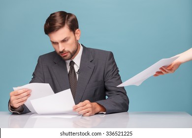 Handsome Male Tv Newscaster Is Preparing For His Report. He Is Reading Documents With Seriousness And Sitting At Desk. The Female Hand Gives Him Papers. Isolated On Blue Background