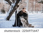 Handsome male warrior with a sword in winter near a snow-covered tree with a wolf. The guy with the husky in the forest in a raincoat. Fantasy image of a prince