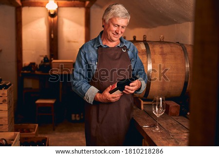 Handsome male winemaker in apron looking at bottle of alcoholic drink and smiling while spending time in wine storage Zdjęcia stock © 