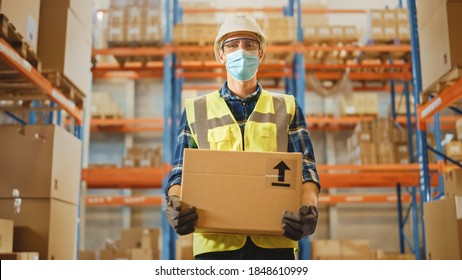 Handsome Male Worker Wearing Medical Face Mask And Hard Hat Carries Cardboard Box Stands In Retail Warehouse Full Of Shelves With Goods. Logistics, Distribution Center. Fromt Shot