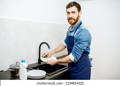 Handsome man in apron doing household chores washing dishes on the kitchen at home - Powered by Shutterstock