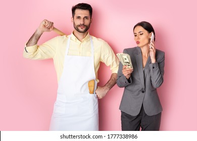 Handsome Man In Apron Holding Wooden Rolling Pin Near Businesswoman With Money Talking On Smartphone On Pink