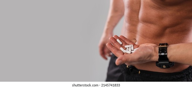 Handsome Man With Athletic Naked Torso Holding Pills In His Hand On Gray Background. Free Space For Text And Design. Healthy Diet And Vitamin Pills. Excitement Pills. 