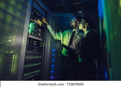 Handsome man and attractive woman are working in data centre with laptop. IT engineer specialists in network server room. Running diagnostics and maintenance. Technicians examining server.