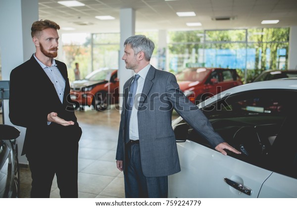 Handsome man at auto\
showroom talking with vehicle dealer. Car sale, sales manager\
showing cars to a\
buyer.