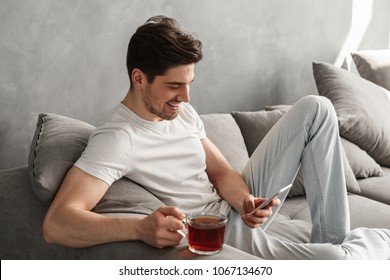 Handsome Man In Basic T-shirt Smiling And Holding Mobile Phone In Hands While Drinking Tea On Couch In House