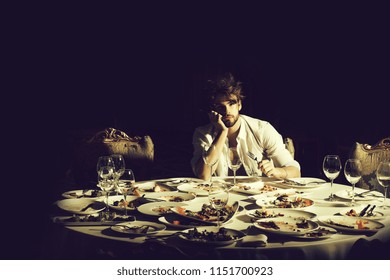 Handsome Man With Beard And Blond Messy Hair Bored Or Tired At Table With Leftovers Or Residues Food On Dirty Plates After Banquet Dinner In Restaurant On Dark Background.