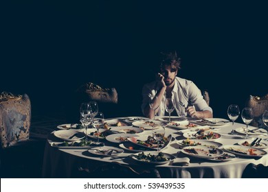 Handsome Man With Beard And Blond Messy Hair Bored Or Tired At Table With Leftovers Or Residues Food On Dirty Plates After Banquet Dinner In Restaurant On Dark Background