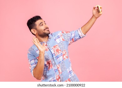 Handsome Man With Beard In Blue Casual Style Shirt Talking On Video Call And Waving Hello Gesture, Having Online Conversation On Mobile Phone. Indoor Studio Shot Isolated On Pink Background.