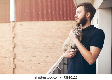 Handsome Man With Beard In Casual Clothes Is Standing On A Terrace And Holding His Beautiful Cat. A Guy With A Cat Is Enjoying Being Outside In The Fresh Air And Sun. Copy-space For Your Advertisement