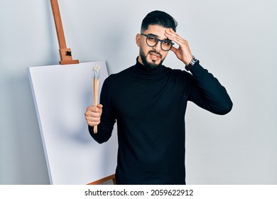 Handsome man with beard holding brushes close to easel stand worried and stressed about a problem with hand on forehead, nervous and anxious for crisis 
