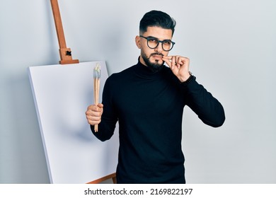 Handsome man with beard holding brushes close to easel stand mouth and lips shut as zip with fingers. secret and silent, taboo talking 