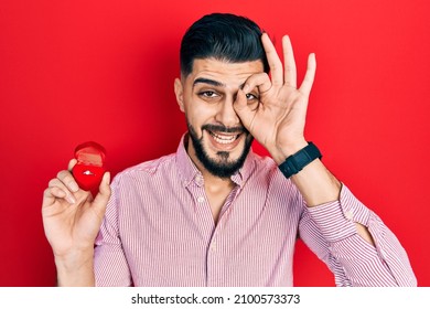 Handsome Man With Beard Holding Engagement Ring For Proposal Smiling Happy Doing Ok Sign With Hand On Eye Looking Through Fingers 