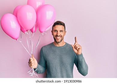 Handsome man with beard holding pink balloons surprised with an idea or question pointing finger with happy face, number one 