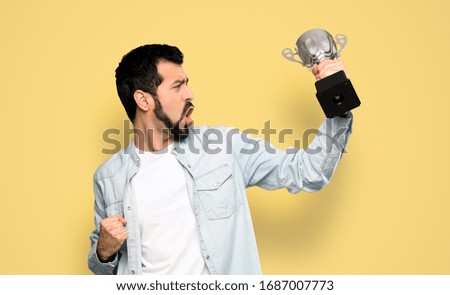 Handsome man with beard holding a trophy over isolated yellow background