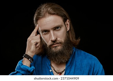 A handsome man with a beard and a medium length haircut in a natural cotton shirt on a black studio background is in his thoughts. People, feelings and emotions.
