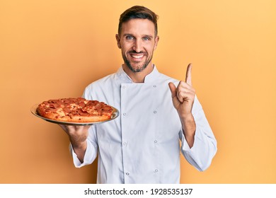 Handsome man with beard professional cook holding italian pizza smiling happy pointing with hand and finger to the side 
