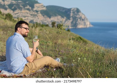 Handsome man with a beard in a shirt and trousers in nature enjoys the view, sits smiling and drinking white wine from a glass. Nature mountains sea. Concept: summer picnic with sea view 