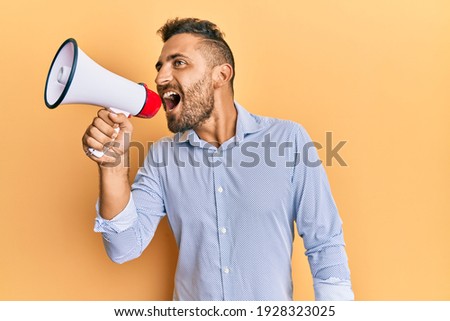 Handsome man with beard shouting through megaphone angry and mad screaming frustrated and furious, shouting with anger. rage and aggressive concept. 