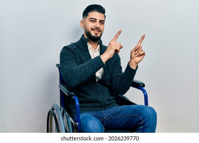 Handsome man with beard sitting on wheelchair smiling and looking at the camera pointing with two hands and fingers to the side. 