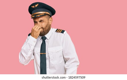 Handsome Man With Beard Wearing Airplane Pilot Uniform Smelling Something Stinky And Disgusting, Intolerable Smell, Holding Breath With Fingers On Nose. Bad Smell 