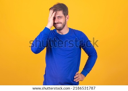 Handsome man with beard, wearing blue shirt over yellow background, putting a hand on his head smiling as if he forgot something