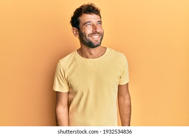 Handsome man with beard wearing casual yellow tshirt over yellow background looking away to side with smile on face, natural expression. laughing confident. 