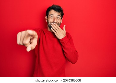 Handsome Man With Beard Wearing Casual Red Sweater Laughing At You, Pointing Finger To The Camera With Hand Over Mouth, Shame Expression 