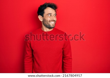 Handsome man with beard wearing casual red sweater looking away to side with smile on face, natural expression. laughing confident. 
