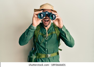 Handsome Man With Beard Wearing Explorer Hat Looking Through Binoculars Smiling And Laughing Hard Out Loud Because Funny Crazy Joke. 