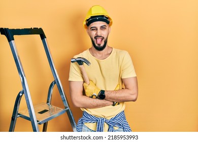 Handsome man with beard wearing hardhat holding hammer by stairs sticking tongue out happy with funny expression. 