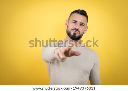 Handsome man with beard wearing sweater over yellow background pointing with finger to the camera and to you, conf