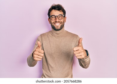 Handsome man with beard wearing turtleneck sweater and glasses success sign doing positive gesture with hand, thumbs up smiling and happy. cheerful expression and winner gesture.  - Shutterstock ID 2057668286