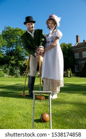 Handsome man and beautiful woman dressed in vintage clothing on lawn in front of stately home, playing croquet with blue sky