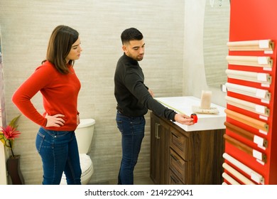 Handsome Man And Beautiful Woman Using A Tape Measure On A Bathroom Cabinet To Remodel Their House At The Shop