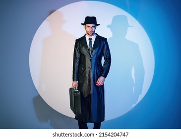 Handsome Man In Black Coat And Hat Holding Briefcase Posing In The Spotlight On Studio Background. Noir Film Style. Private Detective, Spy, Investigation Concept.