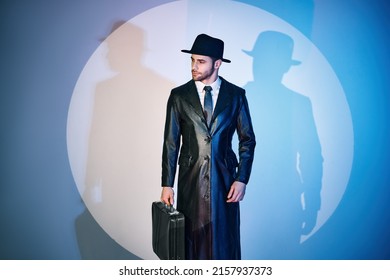 Handsome man in black coat and hat holding briefcase posing in the spotlight on studio background. noir film style. Private detective, spy, investigation concept. 