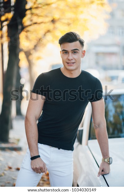 handsome man\
in black shirt and white pants standing in the street leaning on\
the car, on a green background on the land the leaves of the yellow\
autumn time of the year. Fashion\
look
