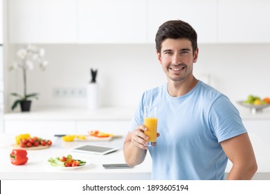 Handsome Man In A Blue T-shirt With A Glass Of Orange Juice In His Hands In The Kitchen At Home, Healthy Lifestyle.