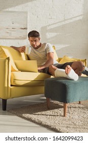 Handsome Man With Broken Leg Using Laptop And Holding Cup On Sofa